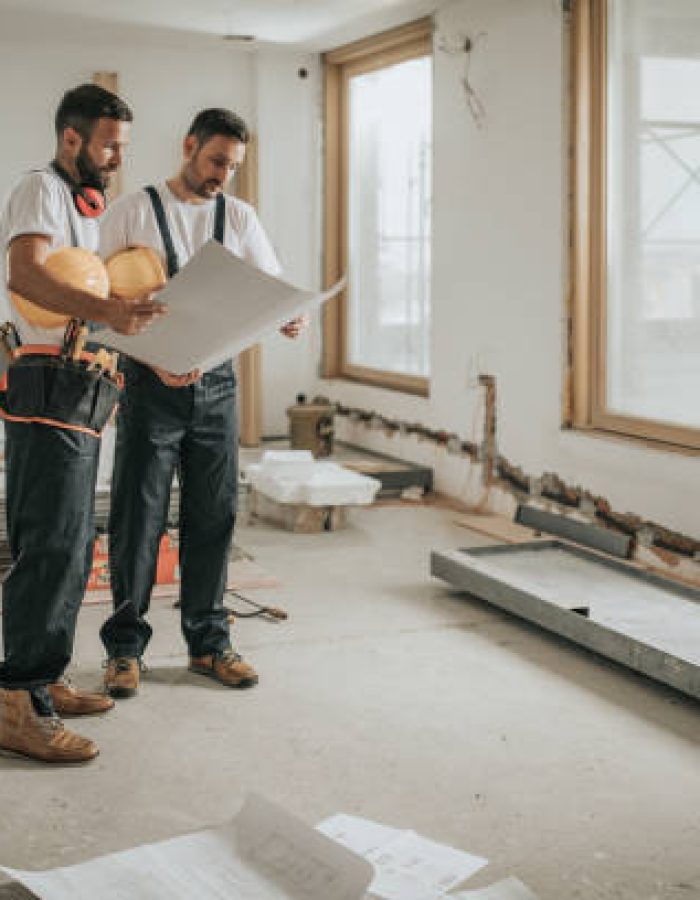 Young manual workers standing at construction site and examining blueprints.